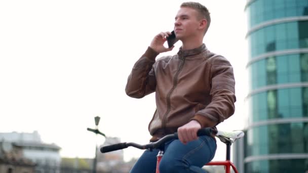 young male bicyclist talks on the phone in downtown slow motion - Materiał filmowy, wideo