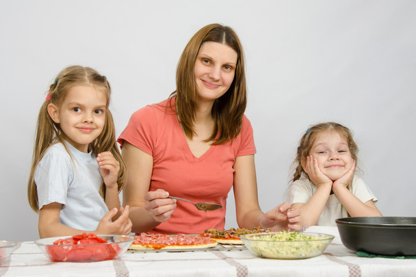 Mama z dwiema małymi dziewczynami siedzi przy stole kuchennym przygotowując pizzę - Zdjęcie, obraz