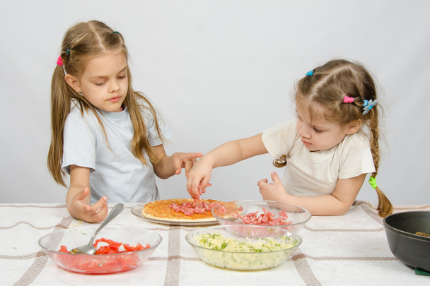 Шестилетняя девочка наблюдает и контролирует свою младшую сестру кладет на пиццу ингредиенты
 - Фото, изображение