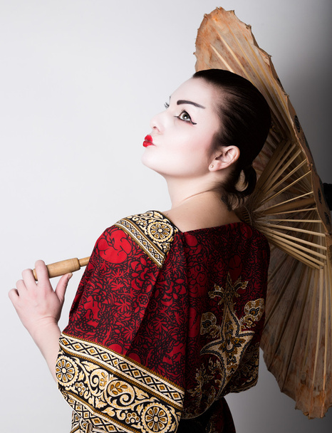 beautiful girl dressed as a geisha girl holding a Chinese umbrella. Geisha makeup and hair dressed in a kimono. The concept of traditional Japanese values - Foto, Imagen