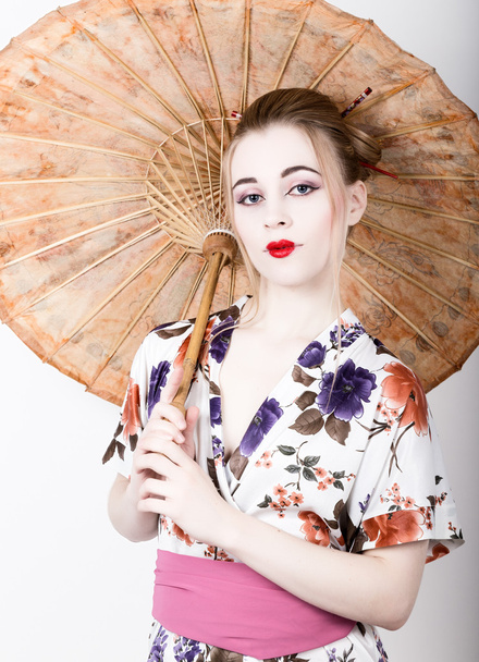 beautiful girl dressed as a geisha girl holding a Chinese umbrella. Geisha makeup and hair dressed in a kimono. The concept of traditional Japanese values - Fotoğraf, Görsel
