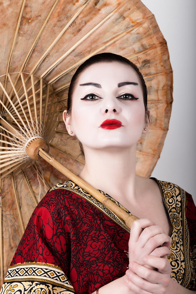 beautiful girl dressed as a geisha girl holding a Chinese umbrella. Geisha makeup and hair dressed in a kimono. The concept of traditional Japanese values - Photo, Image
