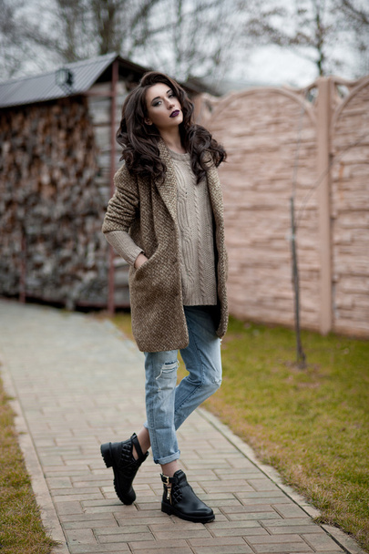 カントリー スタイルを春します。白いニットのセーターと毛皮のような帽子立っている身に着けているセーター カジュアルなの家の近くの庭で素朴な木製の壁の前で若い女性の完全な長さの肖像画. - 写真・画像