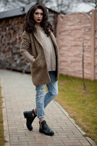 Frühling im Landhausstil. Ganzkörperporträt einer jungen Frau in weißem Strickpullover und Pelzmütze, die vor einer rustikalen Holzwand im Hof neben dem Haus steht und einen lässigen Pullover trägt. - Foto, Bild