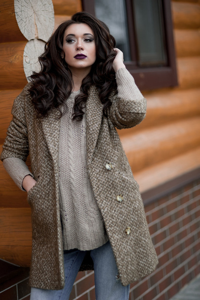 カントリー スタイルを春します。白いニットのセーターと毛皮のような帽子立っている身に着けているセーター カジュアルなの家の近くの庭で素朴な木製の壁の前で若い女性の完全な長さの肖像画. - 写真・画像