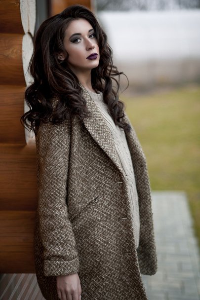 Jaro ve venkovském stylu. Celovečerní portrét mladé ženy v bílém pletený svetr a kožený klobouk stojící před rustikální stěna v zahradě u domu, příležitostné nošení svetr. - Fotografie, Obrázek