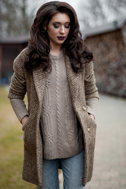 Spring in the country style. Full length portrait of young woman in white knitted sweater and furry hat standing in the front of rustic wood wall in the yard near house, wearing sweater casual. - Photo, Image