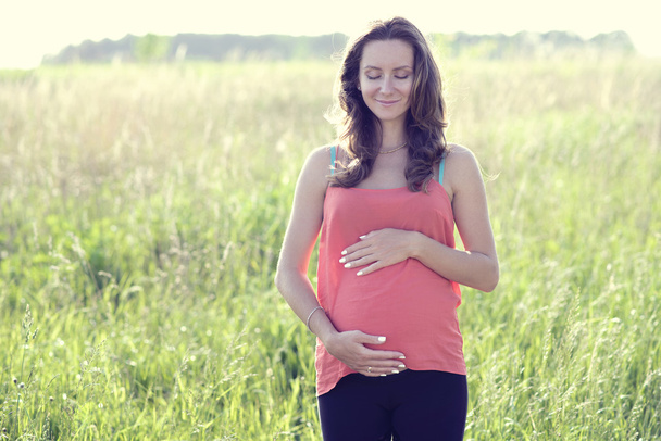 Молода красива вагітна жінка тримає животик посміхаючись, в червоному світле літнє плаття, щаслива на лузі трава
 - Фото, зображення