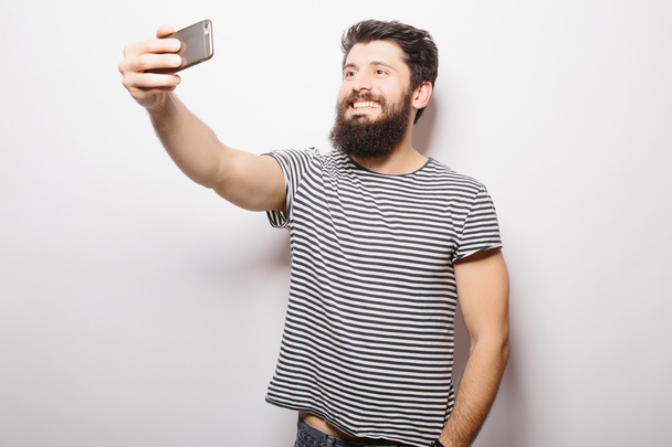 Smiling man with beard standing on white isolated background and taking selfie with mobile phone. - Photo, Image