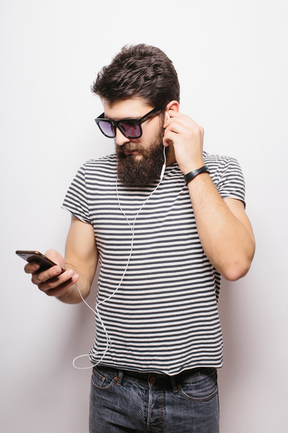 Happy hipster met baard dragen shirt en zonnebril genieten van muziek tegen witte achtergrond - Foto, afbeelding
