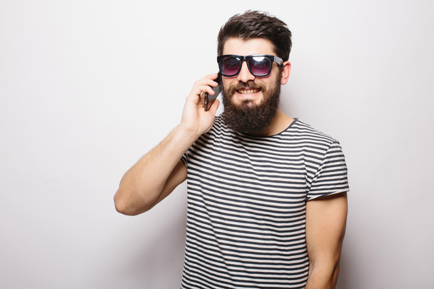 Όμορφος ευτυχισμένος άντρας με γένια στα γυαλιά ηλίου μιλάει στο τηλέφωνο με λευκό φόντο - Φωτογραφία, εικόνα