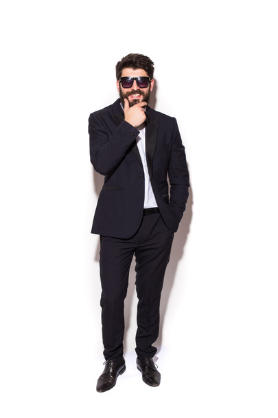 Full height Cheerful young handsome man in sunglasses keeping hand on chin and looking at camera with smile while standing against white background - Valokuva, kuva