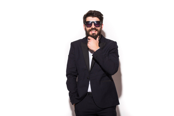 Cheerful young handsome man in sunglasses keeping hand on chin and looking at camera with smile while standing against white background - Foto, Bild