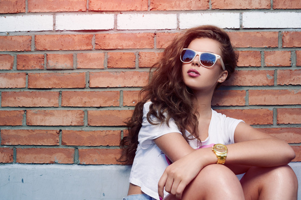 Menina bonita em óculos de sol em um fundo de uma parede de tijolo. Belo cabelo saudável. Calções de ganga
 - Foto, Imagem
