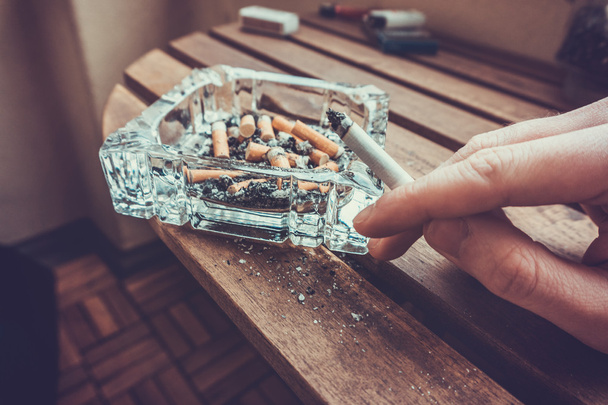 Hombre fumando un cigarrillo sosteniendo una punta de filtro ardiente en su mano junto a un cenicero de vidrio lleno de cenizas y colillas muertas en un concepto de adicción fumador pesado
 - Foto, Imagen
