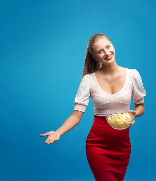 Portrait of fashionable young blond woman in pink-red dress, bright make-up holding, eating fried potato, chips and  over blue background. Copy-space. Unhealthy eating. Junk food concept. - Foto, imagen