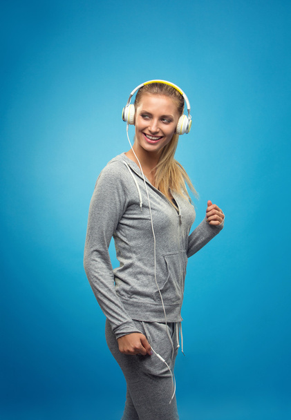 Beautiful blond fit caucasian lady in grey sport  with headphones listening music. Isolated on blue background. - Zdjęcie, obraz