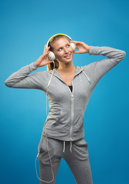 Beautiful blond fit caucasian lady in grey sport  with headphones listening music. Isolated on blue background. - Foto, Imagen
