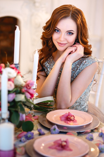 Beautiful young lady in a luxury blue dress sitting by a table with a bouquet of flowers - Foto, Imagen