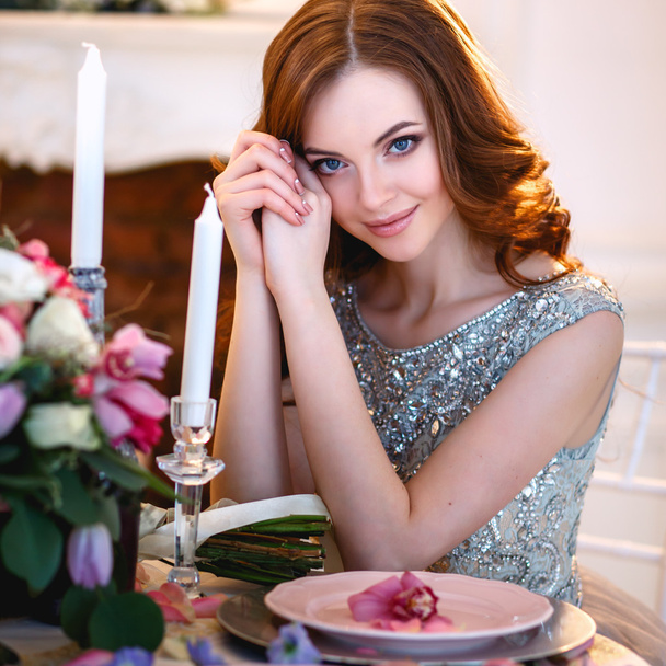 Beautiful young lady in a luxury blue dress sitting by a table with a bouquet of flowers - Photo, Image