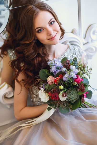 Beautiful young lady in a luxury dress in elegant interior with a bouquet of flowers sitting near mirror - Foto, Imagem