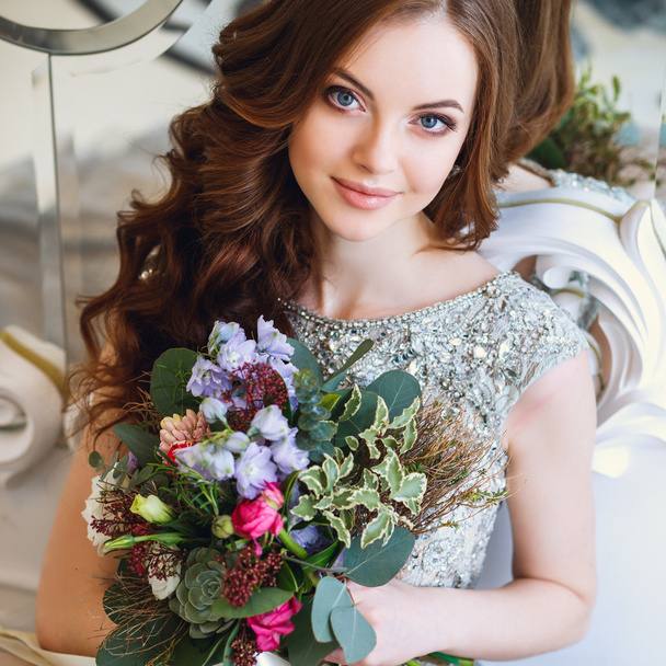 Beautiful young lady in a luxury dress in elegant interior with a bouquet of flowers sitting near mirror - Фото, изображение