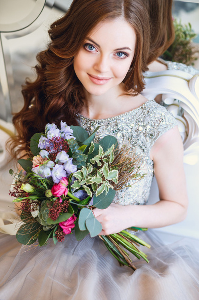 Beautiful young lady in a luxury dress in elegant interior with a bouquet of flowers sitting near mirror - Φωτογραφία, εικόνα