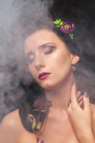 Κομψή, σαγηνευτική μελαχρινή σε ένα σύννεφο καπνού. Μελαχρινή σε ένα όμορφο στεφάνι πάνω σε μαύρο φόντο. - Φωτογραφία, εικόνα