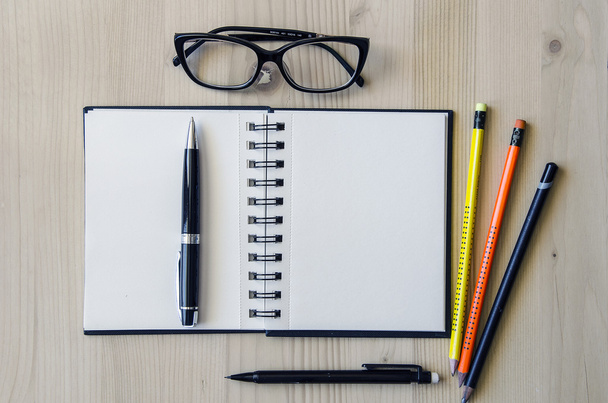 Pluma representativa colocada en un cuaderno en blanco con lápices y gafas sobre un fondo de escritorio de madera. Vista superior
 - Foto, Imagen