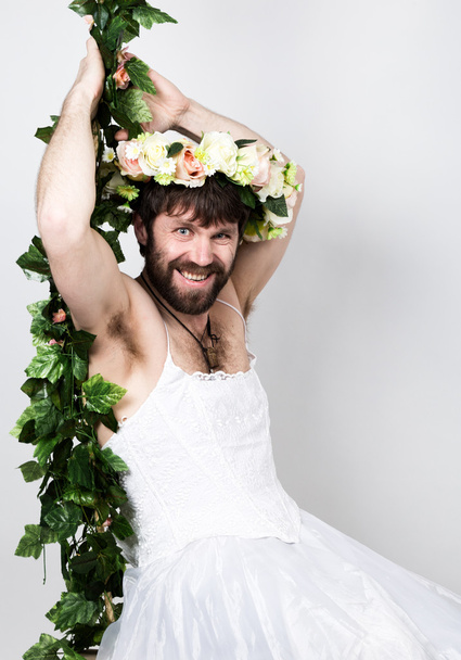 bearded man in a womans wedding dress on her naked body, clinging to the vine. on his head a wreath of flowers. funny bearded bride. woman without hair removal, the natural growth of hair on the body - Fotografie, Obrázek