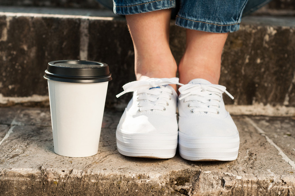 Woman feet in white sneakers standing near a coffee cup - Фото, изображение