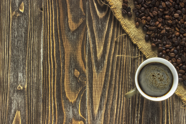 Uma xícara de café e crema está no canto de uma mesa de madeira em uma tela de serapilheira perto de grãos de café desmoronar. Fecha. Vista superior
 - Foto, Imagem
