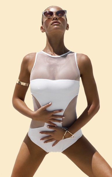 Woman in white swimsuit and golden sunglasses with hair up poses on isolated background. High fashion shot. - Fotó, kép