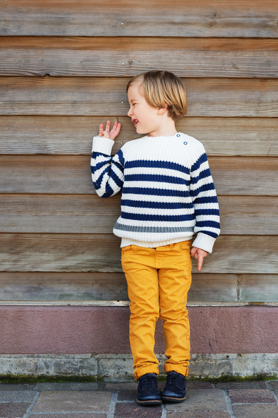 Retrato vertical de moda de niño adorable de 4-5 años con jersey blanco cálido con rayas azules y pantalones amarillos, de pie sobre fondo de madera
 - Foto, Imagen