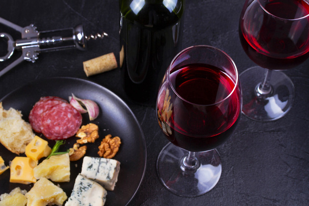 Glass and bottle of red wine, cheese, bread, garlic, nuts, salami on gray stone texture background - Photo, image