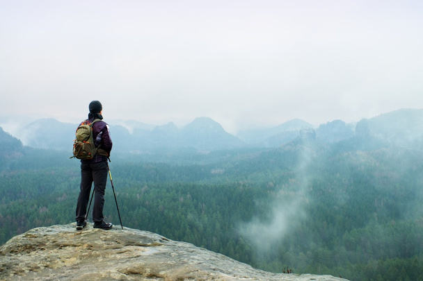 Hiker with backpack and poles on cliff rock  watching over the misty and foggy spring valley to Horizon. - Photo, Image