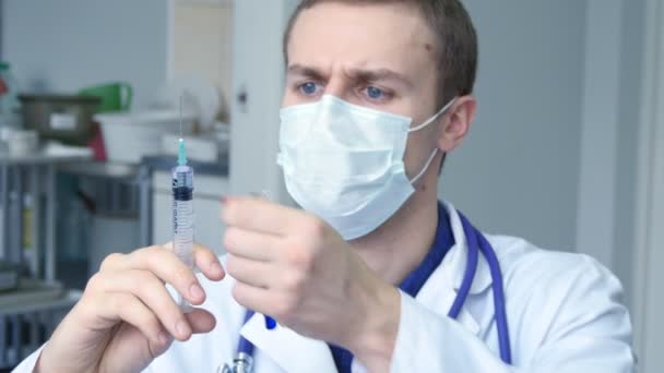 Portrait of young male caucasian doctor pouring a liquid from a syringe to a tube, looking at blue liquid in a test tube and taking off his protective mask - Footage, Video