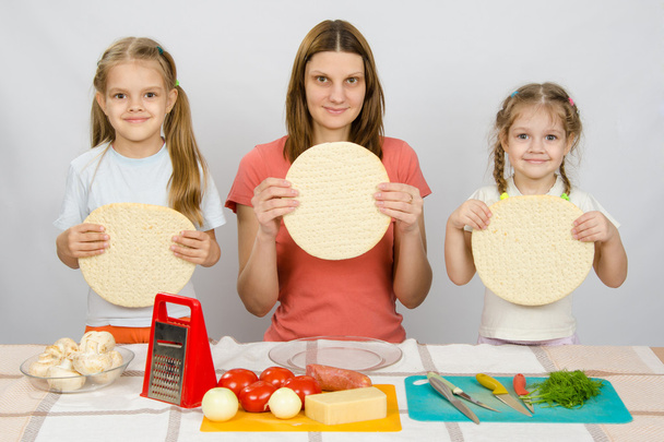 Mama met twee kleine meisjes zittend in een rij aan de keuken tafel en een hand-held pizza bases - Foto, afbeelding