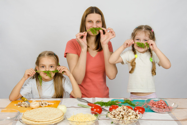 Young housewife with two daughters having fun holding sprig of parsley as a mustache at the kitchen table when sharing cooking - Photo, Image