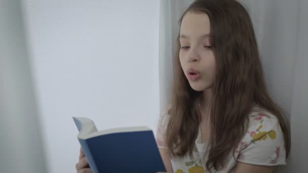 Emotional little girl reading an interesting book by window. - Кадры, видео