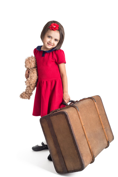 Красивая маленькая девочка улыбается в красном платье с чемоданом и игрушечным медведем
  - Фото, изображение