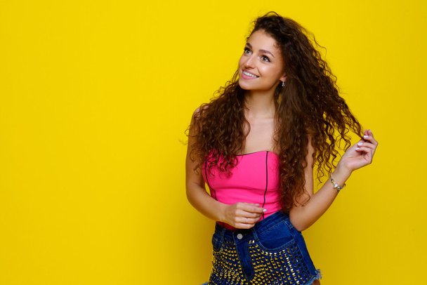 Young and beautiful curly girl in a pink shirt and blue shorts on a yellow background - Foto, afbeelding