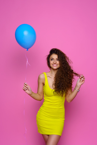 Beautiful and young girl in a yellow dress holding a blue balloon and smiling on a pink background - Foto, Imagem