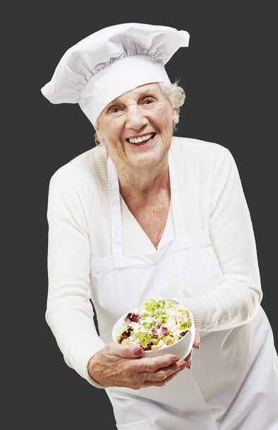 Mulher sênior cozinheiro segurando uma tigela com salada contra uma parte traseira preta
 - Foto, Imagem