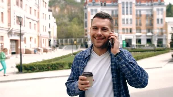 Portret van stijlvolle knappe jongeman met borstelharen permanent buitenshuis. Glimlachende man praten op mobiele telefoon en houden van kopje koffie - Video