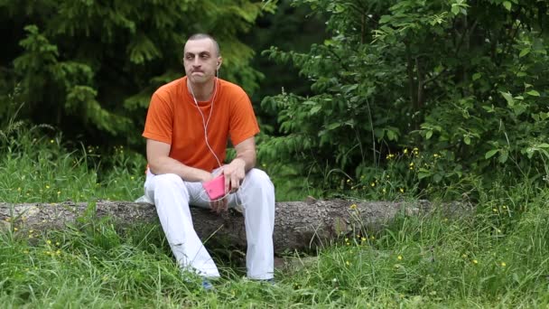 Człowiek z czerwonym smartphone siedzi na spasionego drzewa w lesie i słucha muzyki - Materiał filmowy, wideo