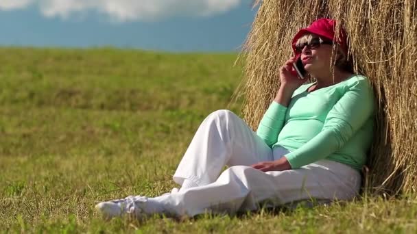 Senior kobieta siedzi w pobliżu stogu siana i komunikuje się za pośrednictwem telefonu komórkowego - Materiał filmowy, wideo