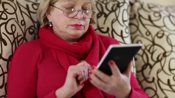 Mujer mayor se sienta en un sofá y utiliza el libro electrónico
.  - Metraje, vídeo
