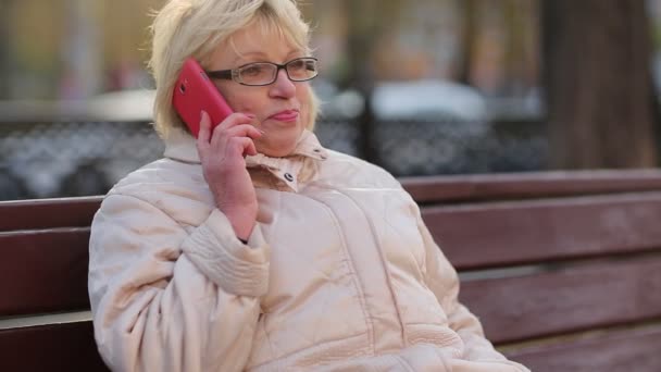 Mujer se sienta en el banco y habla en el teléfono inteligente rojo
 - Metraje, vídeo