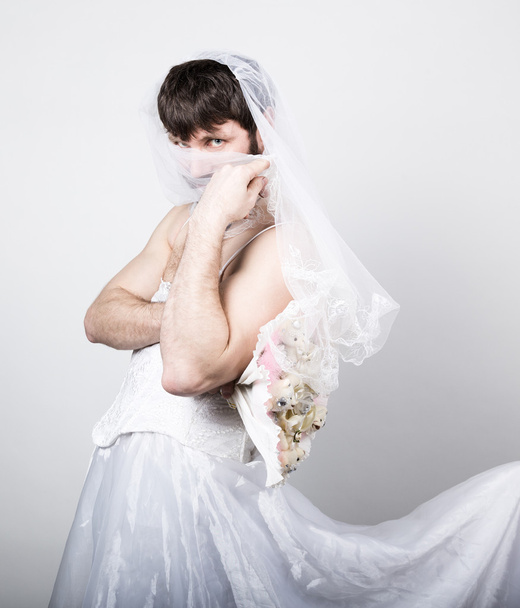 bearded man in a womans wedding dress on her naked body, holding a flower. on his head a veil. funny bearded bride, different emotions - Фото, изображение